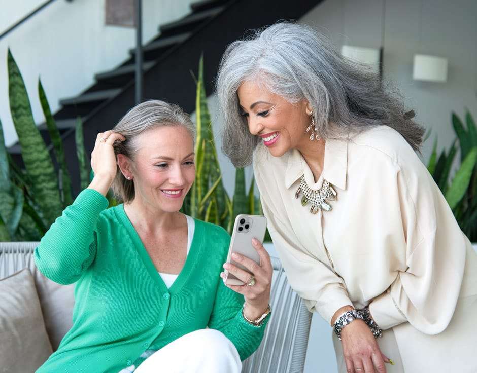 Duas senhoras sorrindo enquanto olham o celular no sofá de casa